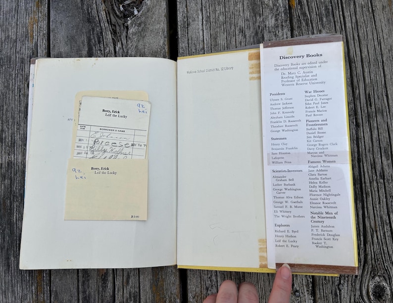 Pu&ograve; includere: Un libro con copertina rigida aperto con una scheda di prestito bibliotecario color crema in una tasca sulla pagina sinistra. La pagina destra presenta un elenco stampato intitolato Discovery Books, che dettaglia varie figure storiche.