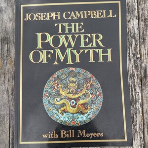 K&ouml;nnte beinhalten: Ein schwarzes Buch mit dem Titel The Power of Myth von Joseph Campbell mit Bill Moyers. Das Cover zeigt goldenen Text und eine kreisf&ouml;rmige, farbenfrohe Illustration eines Drachen, umgeben von komplexen Mustern in Rot, Blau und Gelb.