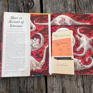 Pode incluir: Um livro de capa dura aberto intitulado Quiet on Account of Dinosaur de Jane Thayer. A p&aacute;gina esquerda apresenta texto impresso, enquanto a p&aacute;gina direita exibe um fundo vermelho com ilustra&ccedil;&otilde;es de dinossauros e um bolso de ficha de biblioteca.