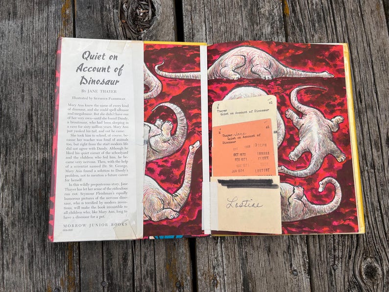 Pode incluir: Um livro de capa dura aberto intitulado Quiet on Account of Dinosaur de Jane Thayer. A p&aacute;gina esquerda apresenta texto impresso, enquanto a p&aacute;gina direita exibe um fundo vermelho com ilustra&ccedil;&otilde;es de dinossauros e um bolso de ficha de biblioteca.