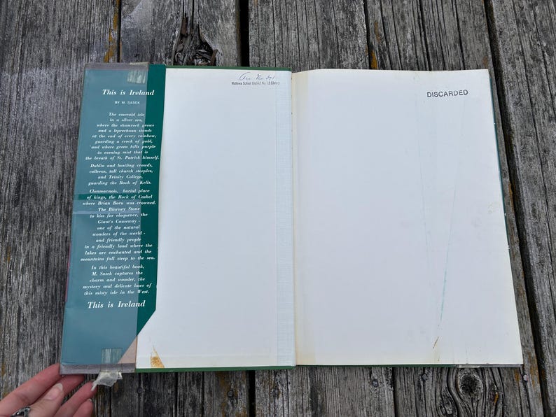 Peut inclure: Un livre reli&eacute; ouvert intitul&eacute; This is Ireland par M. Sasek. Le rabat int&eacute;rieur gauche pr&eacute;sente un texte descriptif sur l'Irlande, tandis que la page de droite est vierge avec le mot DISCARDED imprim&eacute; en noir dans le coin sup&eacute;rieur droit.