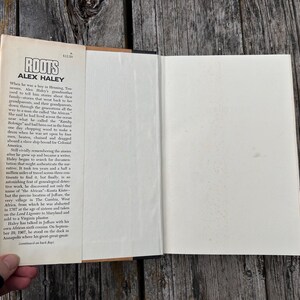 K&ouml;nnte beinhalten: Ein gebundenes Buch mit dem Titel Roots von Alex Haley mit einem cremefarbenen Schutzumschlag. Die vordere Klappe enth&auml;lt schwarz gedruckten Text, der die historische Erz&auml;hlung der Vorfahren des Autors detailliert, inklusive des Preises von 12,50 $.