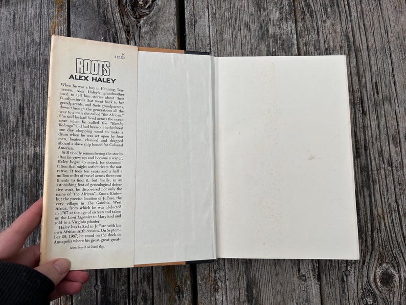 K&ouml;nnte beinhalten: Ein gebundenes Buch mit dem Titel Roots von Alex Haley mit einem cremefarbenen Schutzumschlag. Die vordere Klappe enth&auml;lt schwarz gedruckten Text, der die historische Erz&auml;hlung der Vorfahren des Autors detailliert, inklusive des Preises von 12,50 $.
