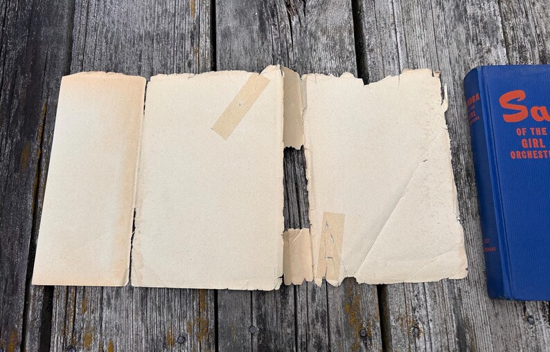 May include: A detached, cream-coloured paper book jacket with frayed edges and visible adhesive tape strips, positioned next to a blue hardcover book with the text "Sally of the Girl Orchestra" printed in orange on the spine.