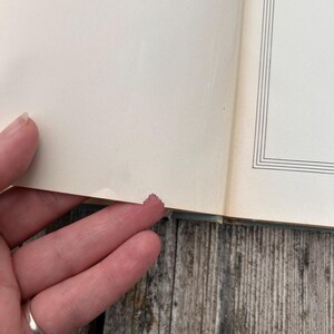 Pu&ograve; includere: Un libro aperto con pagine color bianco sporco che presenta un sottile bordo rettangolare nero sulla pagina destra. Il bordo inferiore della pagina sinistra mostra un piccolo strappo distinto nella carta, con una leggera decolorazione.
