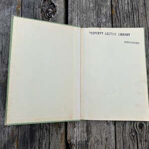 K&ouml;nnte beinhalten: Ein aufgeschlagenes Hardcover-Buch mit hellgr&uuml;nem Einband und cremefarbenen Vorsatzbl&auml;ttern. Auf der rechten Seite befindet sich ein schwarzer Aufdruck mit dem Text PROPERTY LOSTIME LIBRARY sowie dem Wort DISCARDED unter dem Bibliotheksnamen.
