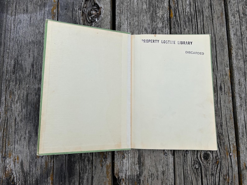 K&ouml;nnte beinhalten: Ein aufgeschlagenes Hardcover-Buch mit hellgr&uuml;nem Einband und cremefarbenen Vorsatzbl&auml;ttern. Auf der rechten Seite befindet sich ein schwarzer Aufdruck mit dem Text PROPERTY LOSTIME LIBRARY sowie dem Wort DISCARDED unter dem Bibliotheksnamen.