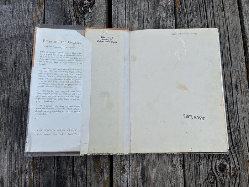 May include: An open hardcover book titled Blaze and the Gypsies by C.W. Anderson, featuring a dust jacket with printed text on the left flap, a cream-colored endpaper with a stamp reading DISCARDED, and a school district property stamp.