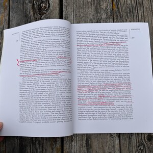 K&ouml;nnte beinhalten: Ein aufgeschlagenes Buch mit wei&szlig;en Seiten und schwarzem Drucktext. Mehrere Textzeilen sind mit roter Tinte markiert, und rote handschriftliche Zeichen befinden sich an den R&auml;ndern. Das Buch liegt flach und zeigt zwei Seiten mit dichtem Text.