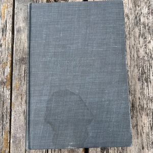 May include: A rectangular hardcover book with a dark gray, textured fabric cover. The front cover features a subtle, embossed script title in the center and a faint, rounded dark stain near the bottom edge, showing a woven material texture.