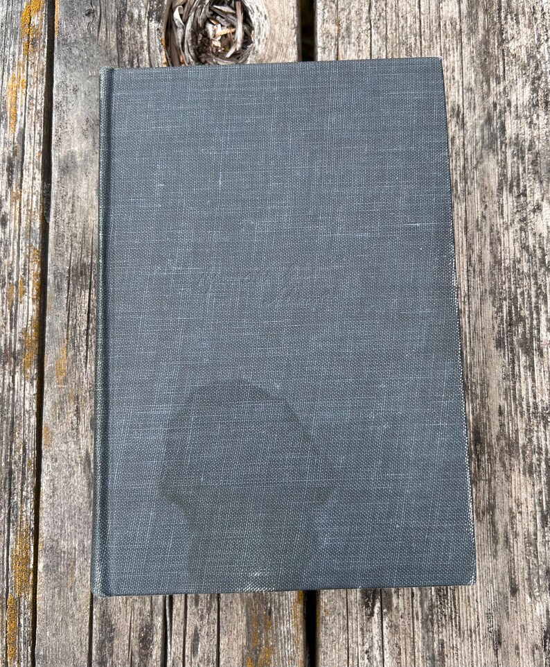 May include: A rectangular hardcover book with a dark gray, textured fabric cover. The front cover features a subtle, embossed script title in the center and a faint, rounded dark stain near the bottom edge, showing a woven material texture.