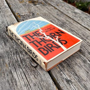 May include: A hardcover book titled The Thorn Birds by Colleen McCullough with a red and white cover featuring a blue sky illustration and a white tree branch graphic. The book has black text on the spine and front cover with visible wear.