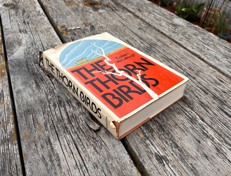 May include: A hardcover book titled The Thorn Birds by Colleen McCullough with a red and white cover featuring a blue sky illustration and a white tree branch graphic. The book has black text on the spine and front cover with visible wear.