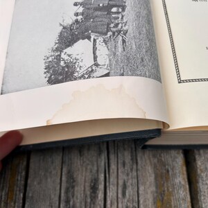 May include: An open hardcover book with a black fabric spine and cream-colored pages. The left page features a black and white historical photograph of people near a tent, with a noticeable light brown water stain along the bottom edge.