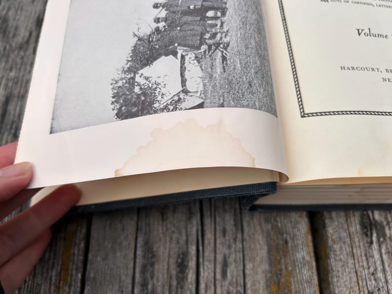 May include: An open hardcover book with a black fabric spine and cream-colored pages. The left page features a black and white historical photograph of people near a tent, with a noticeable light brown water stain along the bottom edge.