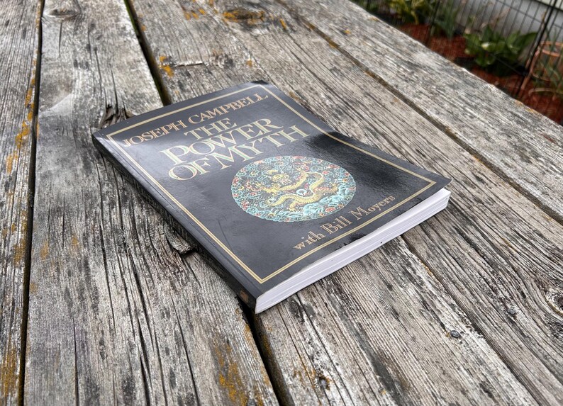 K&ouml;nnte beinhalten: Ein schwarzes Taschenbuch mit dem Titel The Power of Myth von Joseph Campbell mit Bill Moyers. Das Cover zeigt goldenen Text und eine kreisf&ouml;rmige Illustration eines goldenen Drachen auf t&uuml;rkisfarbenem Grund. Das Buch hat wei&szlig;e Seiten und ein gl&auml;nzendes Finish.
