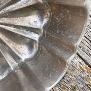 Może przedstawiać: Srebrzysta metalowa forma do pieczenia o karbowanym, falistym wzorze. Wewnętrzna powierzchnia formy ma metaliczne wykończenie z widocznymi śladami użytkowania, w tym jasnymi plamami i teksturą na zakrzywionych, promienistych żebrach.