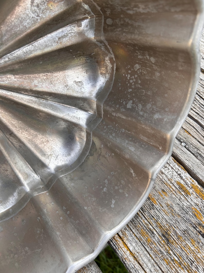 Może przedstawiać: Srebrzysta metalowa forma do pieczenia o karbowanym, falistym wzorze. Wewnętrzna powierzchnia formy ma metaliczne wykończenie z widocznymi śladami użytkowania, w tym jasnymi plamami i teksturą na zakrzywionych, promienistych żebrach.