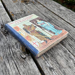 Może przedstawiać: Książka w twardej oprawie zatytułowana King Arthur and the Knights of the Round Table, A Golden Illustrated Classic. Okładka przedstawia ilustrację rycerza w zbroi oraz postaci w niebieskiej szacie, wraz z kilkoma herbami.