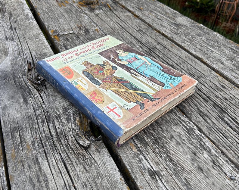 Może przedstawiać: Książka w twardej oprawie zatytułowana King Arthur and the Knights of the Round Table, A Golden Illustrated Classic. Okładka przedstawia ilustrację rycerza w zbroi oraz postaci w niebieskiej szacie, wraz z kilkoma herbami.