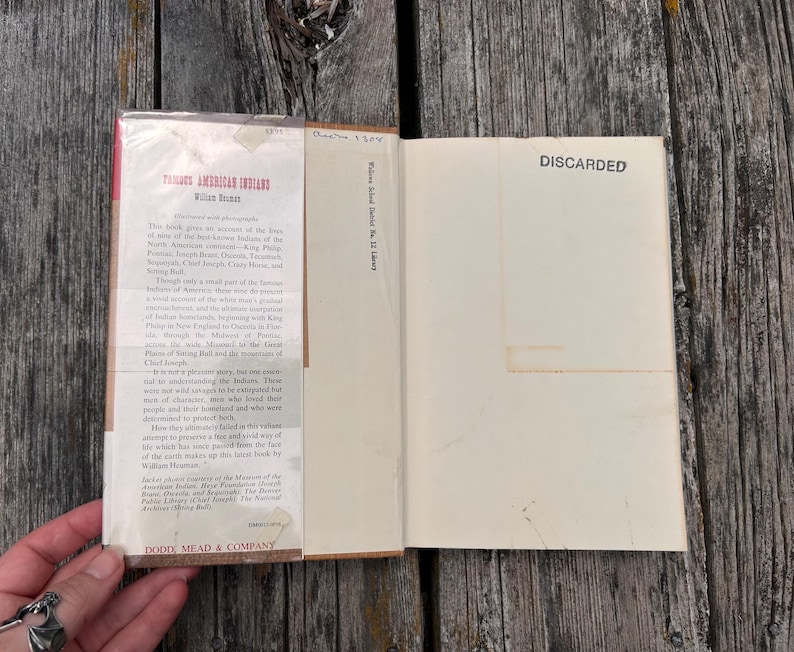 Puede incluir: Un libro de tapa dura titulado Famous American Indians de William Heuman con una sobrecubierta que presenta texto hist&oacute;rico. La p&aacute;gina interior tiene la palabra DISCARDED impresa en tinta negra. El libro est&aacute; abierto, mostrando la solapa.