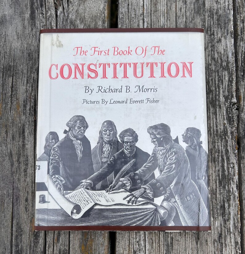 Puede incluir: Un libro de tapa dura titulado The First Book Of The Constitution de Richard B. Morris con ilustraciones de Leonard Everett Fisher. La cubierta blanca presenta una ilustraci&oacute;n en blanco y negro de figuras hist&oacute;ricas firmando.