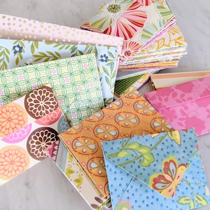 May include: A collection of colorful, patterned paper envelopes in various sizes and designs. The envelopes feature floral, geometric, and polka dot patterns in shades of pink, blue, green, orange, and gold. Some are folded, while others are stacked.