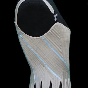 May include: A beige corset with light blue ribbon accents and a scalloped hem. The corset is displayed on a black mannequin, showcasing its structured design and intricate stitching. The shoulder straps are also beige.