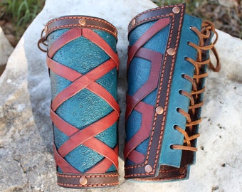 Puede incluir: Un par de protectores de brazo de cuero azul turquesa con correas y cordones de cuero marrón. Las correas están cruzadas y tienen un patrón decorativo. Los protectores están asegurados con remaches de cobre.