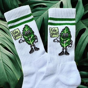 Könnte beinhalten: Weiße Socken mit grünen Streifen und einem Cartoon-Kohl-Charakter, der einen grünen Hut trägt und eine Sprechblase hält, auf der "Kale yeah!" steht.