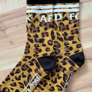 Może przedstawiać: Para czarno-żółtych skarpet z wzorem lamparta. Skarpety mają biały pasek i napis "KAFD FC" na górze. Spód skarpet ma napis "GREEN LIGHT" w kolorze białym.