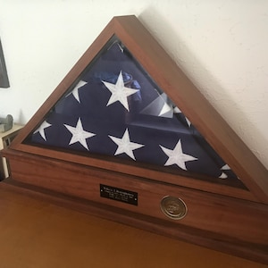 May include: A wooden flag case with a glass front, displaying a folded American flag. The case has a brass plaque with an inscription and a round brass emblem.