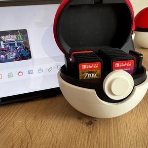 Peut inclure: Un support de cartouches de jeu en forme de Poké Ball rouge et blanc, ouvert pour révéler plusieurs cartouches de jeu. Une console Nintendo Switch est en arrière-plan, affichant des icônes de jeu. D'autres supports Poké Ball sont visibles.