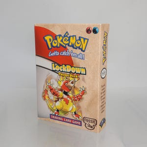 May include: A rectangular Pokémon Fossil Lockdown theme deck trading card game box. The front features the Pokémon Magmar, the Pokémon logo, and the text Gotta catch 'em all! and Lockdown Theme Deck. The box has a tan background with red accents.