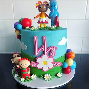 May include: A round cake with light blue and green frosting, decorated with In the Night Garden character figurines, colorful spheres, and white fondant daisies. A pink sign on the front reads Lily is one in white script lettering.