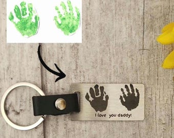 May include: A silver rectangular keychain with two handprints and the text "I love you daddy!". The keychain has a black leather strap and a silver keyring. Above the keychain are two green handprints.