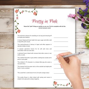 Op de afbeelding: Een wit papieren spelvel met de titel "Pretty in Pink" met invulvragen over roze items. Het vel is versierd met roze bloemaccenten. Een hand met een pen staat klaar om op het vel te schrijven.