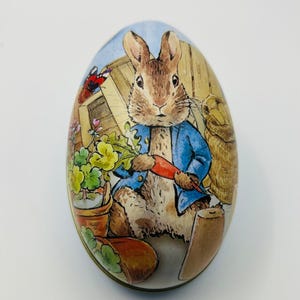 Puede incluir: Una lata redonda con forma de huevo con una ilustración colorida de un conejo sosteniendo una zanahoria. El conejo lleva una chaqueta azul y está rodeado de plantas y una cesta.