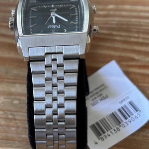 May include: A silver Pulsar watch with a rectangular black face and a metal strap. The watch has a square face with white hands and markers. The brand name "Pulsar" is printed on the watch face.