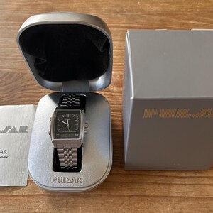 May include: A silver Pulsar watch with a black face and a stainless steel strap, presented in its original case. The watch is alongside a box and a pamphlet, both with the Pulsar logo and text.