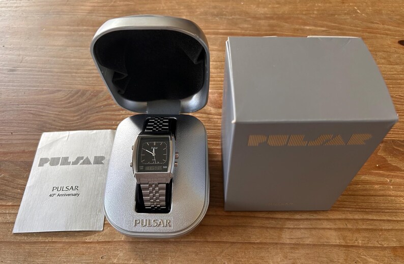 May include: A silver Pulsar watch with a black face and a stainless steel strap, presented in its original case. The watch is alongside a box and a pamphlet, both with the Pulsar logo and text.