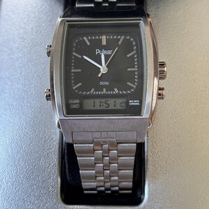 May include: A silver Pulsar watch with a black square face and a digital display showing 11:51:10. The watch has a silver strap and the word "Pulsar" is printed on the watch face and the base. The watch is displayed on a silver stand.