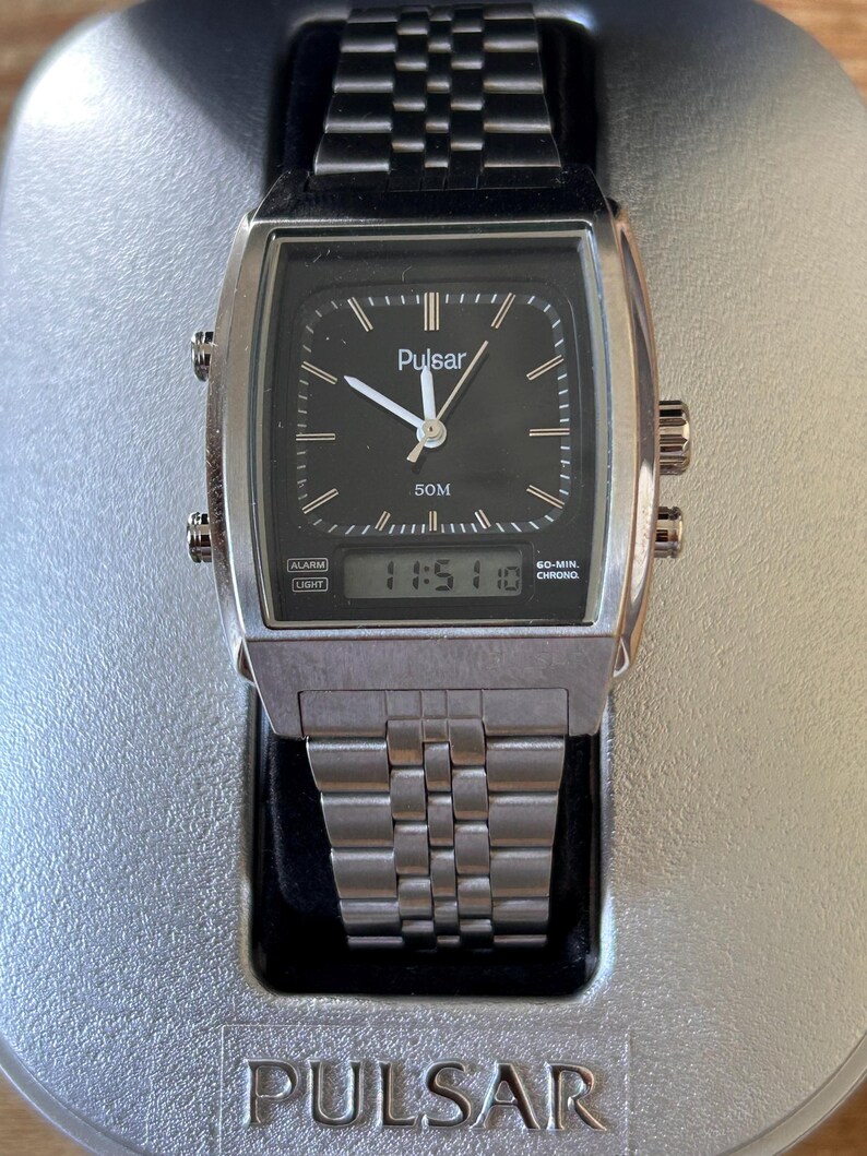 May include: A silver Pulsar watch with a black square face and a digital display showing 11:51:10. The watch has a silver strap and the word "Pulsar" is printed on the watch face and the base. The watch is displayed on a silver stand.