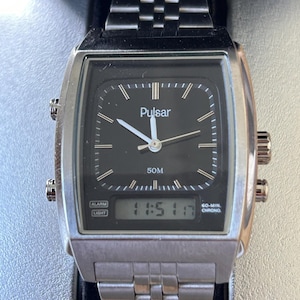May include: Silver Pulsar watch with a black face and a rectangular design. The watch features a digital display showing "11:51 17" and analog hands. The watchband is made of linked metal. The watch has two side buttons.