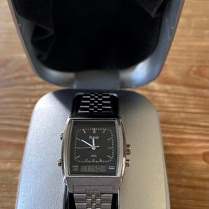May include: A silver Pulsar watch with a black square face and a metal strap, presented in an open silver case. The watch face shows the time as 11:50:49. The case is lined with black fabric, and the brand name is visible.