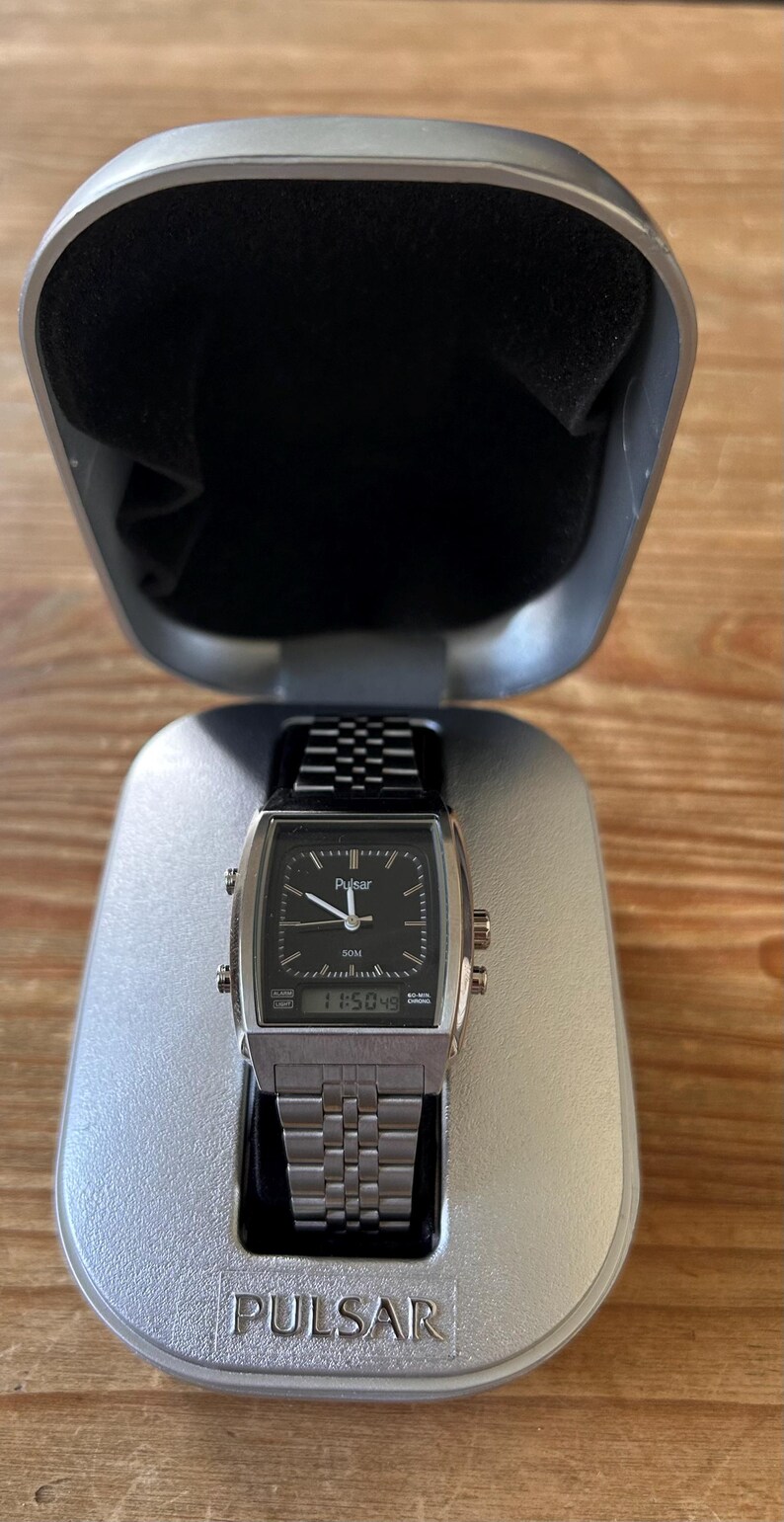 May include: A silver Pulsar watch with a black square face and a metal strap, presented in an open silver case. The watch face shows the time as 11:50:49. The case is lined with black fabric, and the brand name is visible.