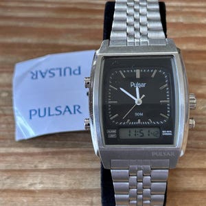 May include: A silver Pulsar watch with a black face and digital display showing 11:51:42. The watch has a rectangular case and a linked metal strap. A white paper with "Pulsar" printed on it is visible.