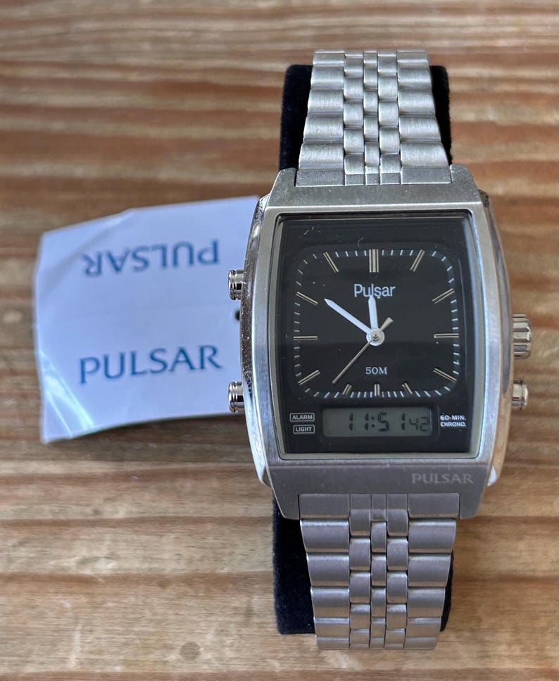 May include: A silver Pulsar watch with a black face and digital display showing 11:51:42. The watch has a rectangular case and a linked metal strap. A white paper with "Pulsar" printed on it is visible.