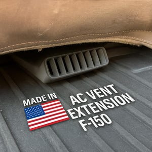 May include: A gray AC vent extension for an F-150 truck, with the text "AC VENT EXTENSION F-150" in white. The image also includes a small American flag and the words "MADE IN".
