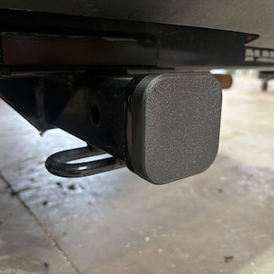 May include: A black trailer hitch receiver with a square, textured, black cover. A U-shaped loop is visible below the receiver. The image is taken from a low angle, showing the underside of a vehicle.